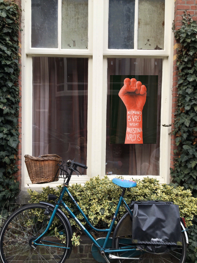 Foto van de voorgevel van een woning, waar een fiets voor geparkeerd staat. Achter de raam zit een poster van een rode vuist tegen een zwarte achtergrond, met daarop de tekst: &lsquo;Niemand is vrij, totdat Palestina vrij is&rsquo;